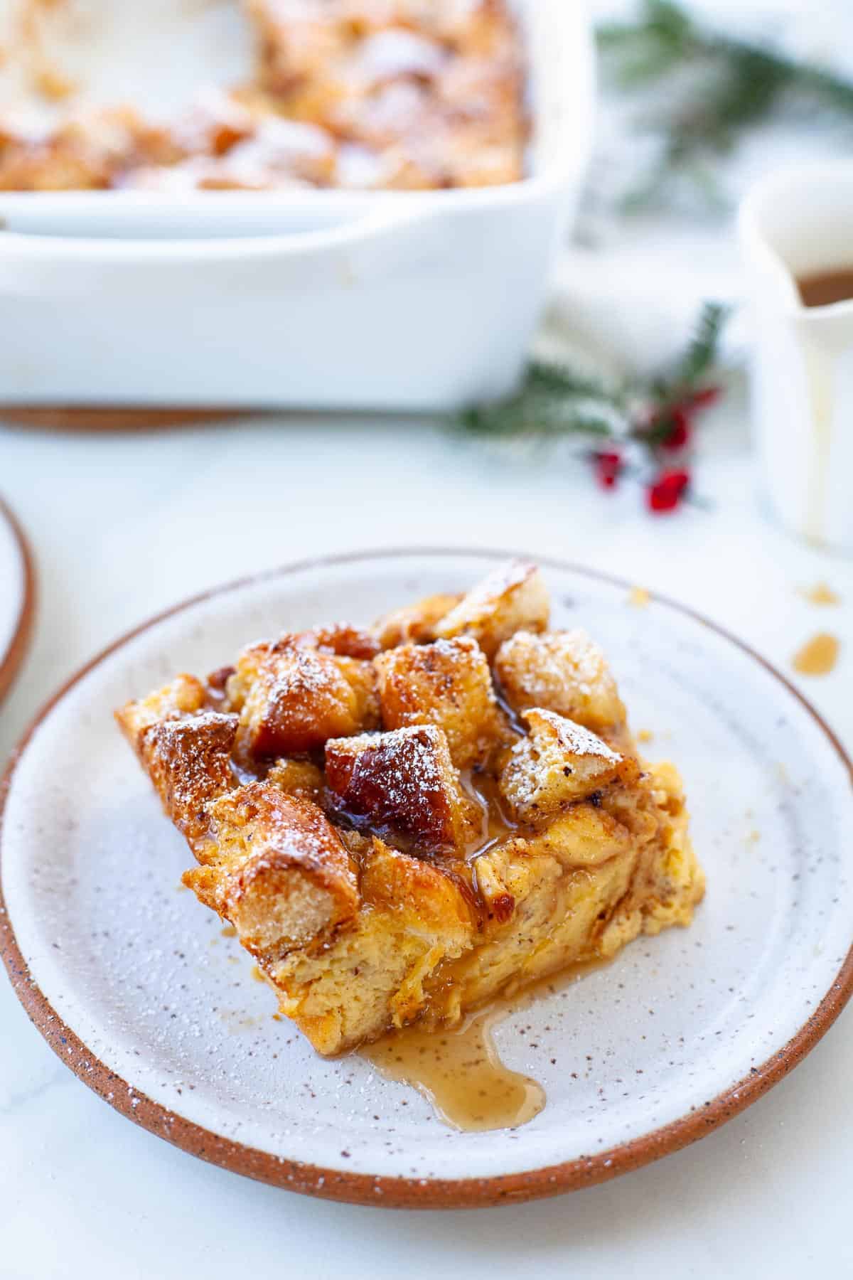 eggnog bread pudding in white speckled plate and white background with Christmas foliage in background 